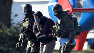 A bearded man, blindfolded, with an earguard and his hands tied in heavy mittens, is escorted by a heavily armed police in full tactical dress, his face concealed by hoods