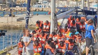 Migrants wearing high-viz lifejackets are seen on the deck of a ship docked in port