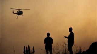 Silhouettes figures watch a helicopter against a sky turned orange from a bushfire at Forster, New South Wales on 7 November
