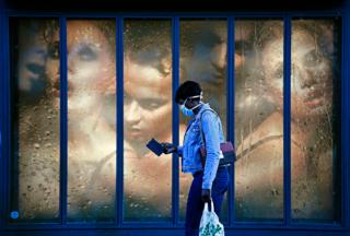 A woman wearing a protective mask walks past a store window with an advert behind her showing lots of women crowded together