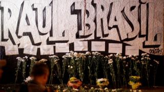 Flores e homenagens em frente a muro grafitado: "Raul Brasil"