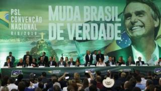 Convenção do PSL tem representantes do partido sentados à mesa sobre palco; no fundo, painel grande com imagem de Bolsonaro e dizeres: 'Muda Brasil de verdade'