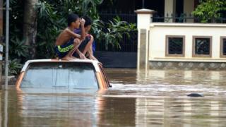 Anak-anak duduk di atas kap mobil di tengah genangan banjir yang melanda satu perumahan di Makassar.