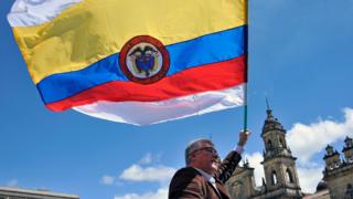Un hombre con una bandera de Colombia con dos franjas más, una arriba y una abajo, de color blanco, símbolo universal de la paz.