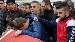 Scene of the funeral of Ayhan Arik in an Istanbul mosque, showing three young males by a coffin, the youngest boy, left, with eyes downcast; in the middle, Ulas, a teenager, head bowed over the coffin; and right, an older boy wearing a beard, one hand on Ulas' shoulder and holding back a tear.