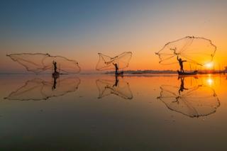 Three fisherman throwing their nets out to sea with a sunset behind them