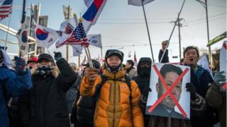 Протестующие держат флаги США и Южной Кореи и вычеркнута вывеска с лицом Ким Чен Ына.