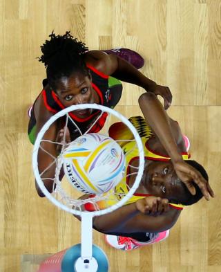 Two Netball players stand next to the net, watching the ball go in. They are both reaching upwards and looking intently at the ball.