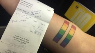 Receipt which says 'Can't tip someone who doesn't love Jesus! Bad tattoo,' placed next to a rainbow equal sign tattoo