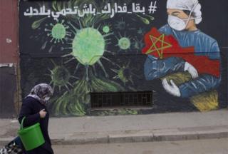 A woman wearing a mask walks past a large mural depicting a masked health worker cradling the nation of Morocco in their arms.