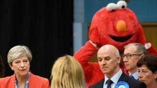 Theresa May participa de divulgação dos resultados da eleição, com homem fantasiado ao fundo