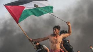 شاب عاري الصدر يحمل العلم الفلسطيني بيمينه ومقلاعا في يساره. صورة بعدسة الصحفي المصور محمد حسونة