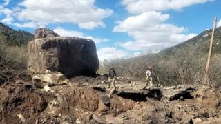 The stretch of road that was destroyed by a rock slide containing two huge rocks with two workers standing in the road.