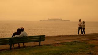 Orange skies across San Francisco caused by wildfires in California, couple sat on beach and another couple walking. Alcatraz is in the distance across the water.