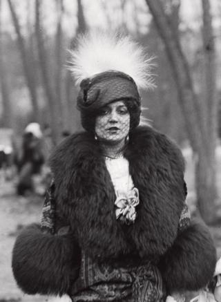 A portrait of a woman in a park wearing an elaborate hat, face veil and thick fur coat