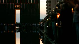 A 09:03 archway and pool lined with people remembering the attack, with candles