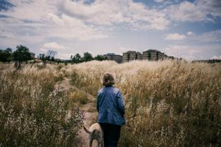 A lady stands in a field with a dog on a lead next to a residential