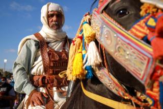 A man dressed in ornate, brightly coloured clothes sits astride a horse whose bridle is similarly adorned.