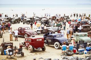 People gather on a beach with vintage cars