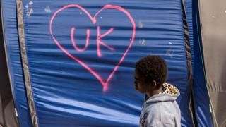 Child walks past a tent in Calais Jungle which has "UK" in a love heart written in graffiti