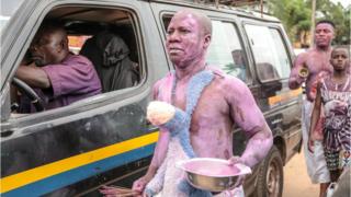 Men covered in purple paint walking next to a car in a street parade in Arondizuogu during the Ikeji Festival in Nigeria
