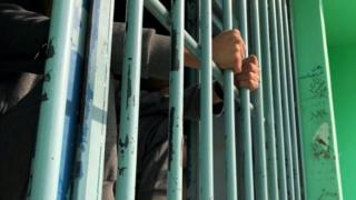 A boy clutches onto the bars of a cell window