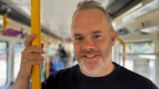 Keith Watson, who has grey hair and a beard and wears a black T-shirt, standing in a train carriage holding onto a yellow pole.