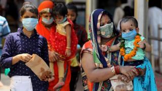 Mulheres com roupas tradicionais indianas usam máscaras contra covid-19 e carregam crianças, também de máscaras, no colo