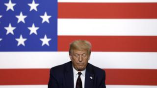 Trump olha para baixo, com bandeira dos EUA ao fundo