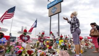 Três pessoas em frente a flores e outras demonstrações de homeangem em área externa do local onde ocorreu ataque em El Paso