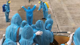 People disembarking from a plane in Vietnam are wearing protective plastic suits and being sprayed with disinfectant