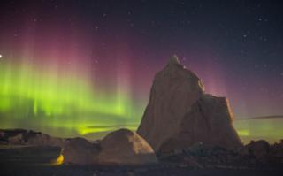 An igloo by an iceberg with the northern lights in the sky. Photograph by Clare Kines