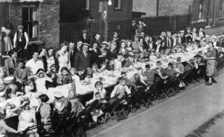 A VE Day celebration street party with people having a meal at a long table