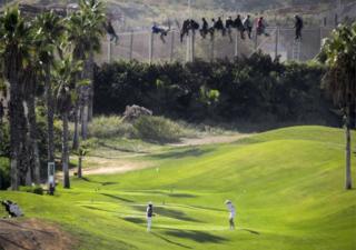 A golfer hits a tee shot as African migrants sit atop a border fence during an attempt to cross into Spanish territories between Morocco and Spain's north African enclave of Melilla