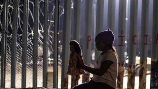Niña migrante con muñeca al lado del muro
