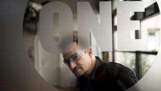 Bono looks through a sheet of glass which is etched with the logo of his charity organisation One