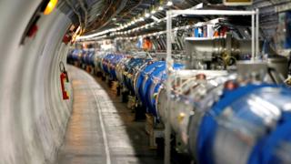 An image shows high-tech machinery in a long array, almost like a very large tube, running through a concrete tunnel, with a staggering array of cables and gleaming industrial machinery connected to the tube