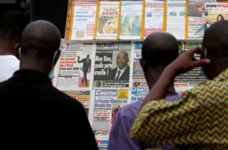 People stand in front of a news stand full of front pages reporting the death of the prime minister.