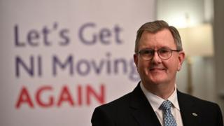 Sir Jeffrey Donaldson in front of a sign that says "Let's Get NI Moving Again"