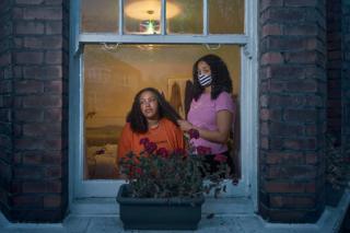 A woman combs a woman's hair next to a window, both wearing masks