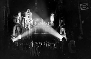 Floodlights illuminate the building tops near St Paul's Cathedral with revellers on the street