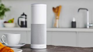 A white Amazon Echo smart speaker stands on a kitchen countertop with staged coffee and croissant