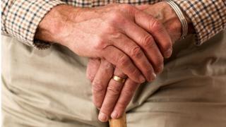 Mãos de um homem idoso, cruzadas uma sob a outra; ele tem uma larga aliança dourada na mão esquerda; veste uma camisa chadrez