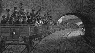 This print shows commuters waving their hats in the air during a trial journey on the London Metropolitan Underground railway.