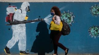 Woman in mask walking past graffitti of person spraying disinfectant on coronavirus images