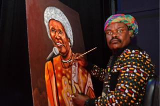 Rasta, whose real name is Lebani Sirenje, sit at an easel painting a large canvas with a brush.