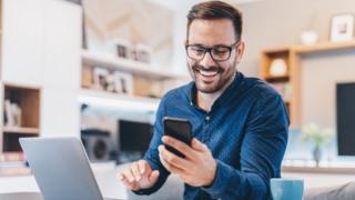 A man laughs at something on his phone while working from home in this photo