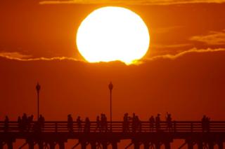 The sun sets behind a cloud as people cool off with a walk along an ocean pier in Oceanside, California, US on 5 July 2018
