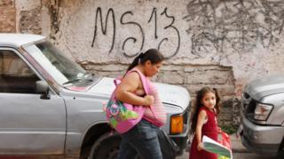 Mujer y niña caminan frente a una pared con la inscripción MS-13
