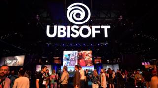Yoğun bir ticaret fuarında havada büyük bir Ubisoft işareti asılı, insanlarla dolu ve vitrin oyunları sergiliyor